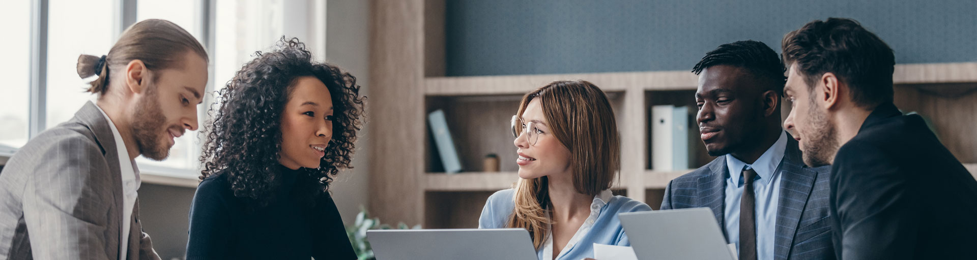Five people wearing business attire sitting around table with laptops
