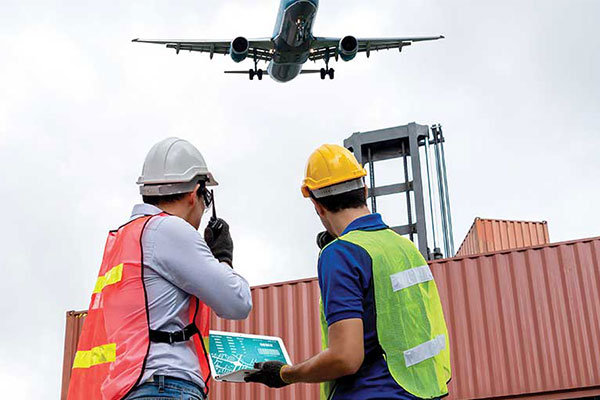 Two construction workers in helmets and vests check paperwork beside stacked shipping containers