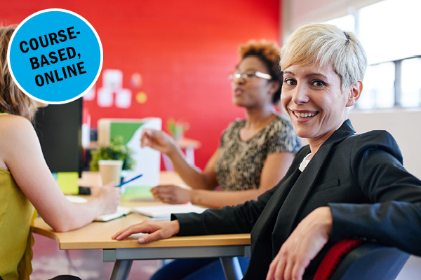 A woman smiles confidently at the camera in a bright room with two colleagues discussing in the background