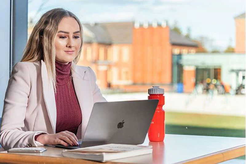 A woman with long blonde hair sits at a desk near a large window, working on a laptop. She is wearing a light pink blazer over a maroon turtleneck, and a red water bottle and a notebook are placed on the desk beside her.