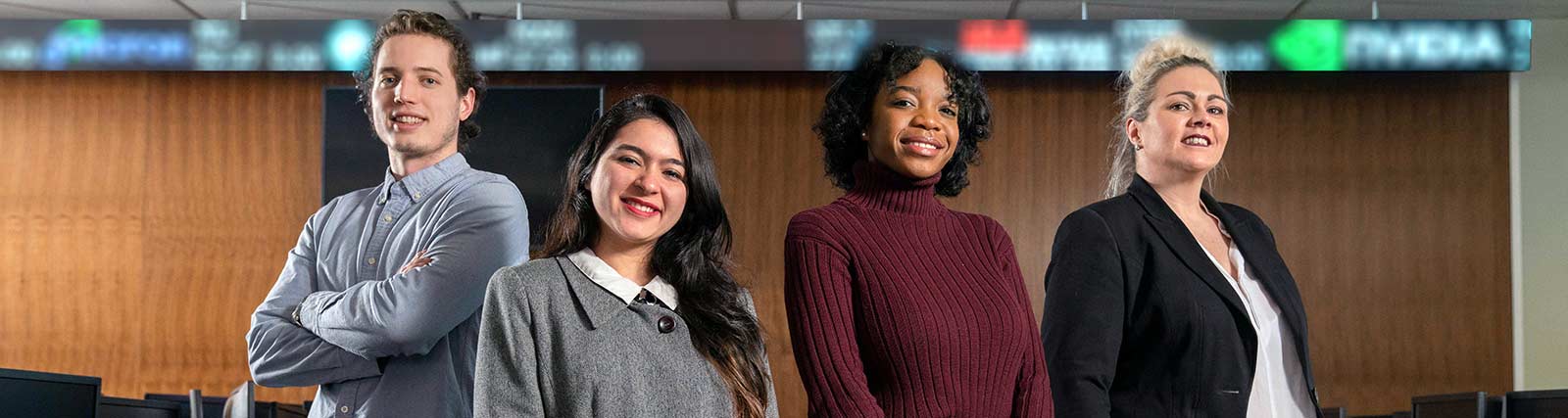 Four people stand confidently in an office with a digital stock ticker above them