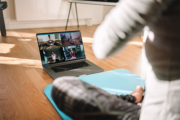 View of a woman conducting virtual fitness class with group of people at home on a video conference. Fitness instructor taking online yoga classes over a video call in laptop.