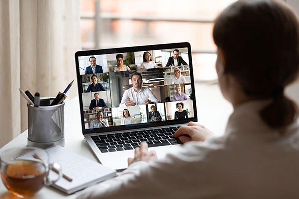 View over businesslady shoulder seated at workplace desk look at computer screen where collage of many diverse people involved at video conference negotiations activity