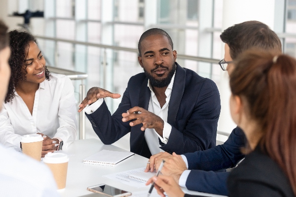 Man dressed in a suit sitting at a table in an office environment and speaking to others