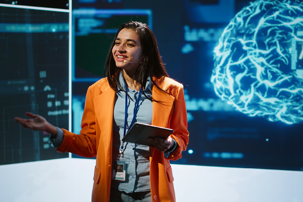 A young professional women wearing a headset and speaking to an audience with large monitors behind her