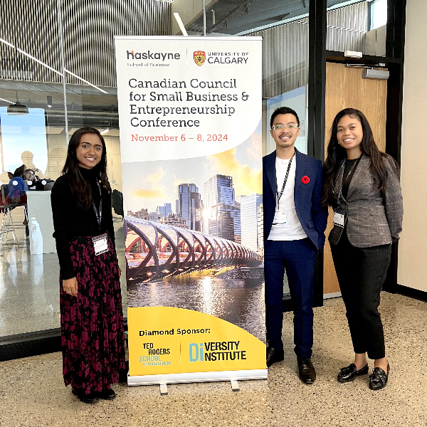 John, Sahana, and Ashley at the CCSBE conference standing in front of a banner