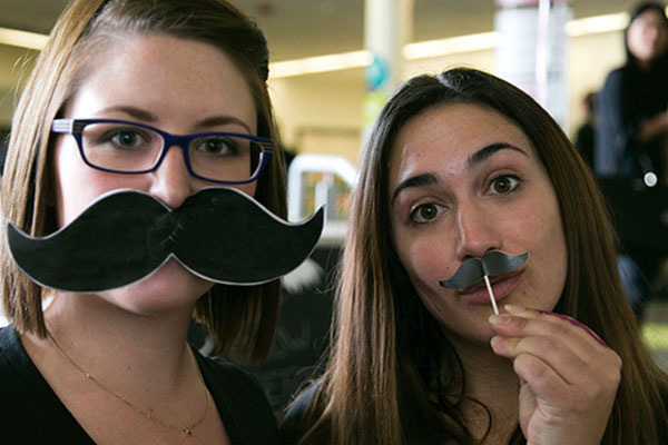 2 female students holding mustache in front of lips