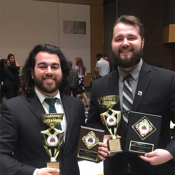 Two male students in suits holding trophies